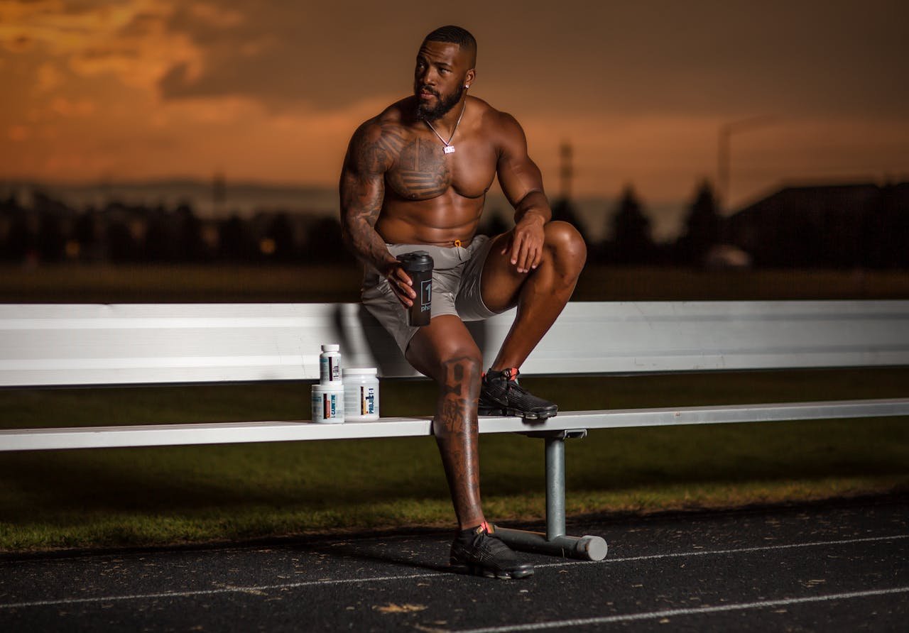 Services Muscular athlete sitting on a bench with supplements, enjoying a sunset at an outdoor stadium.