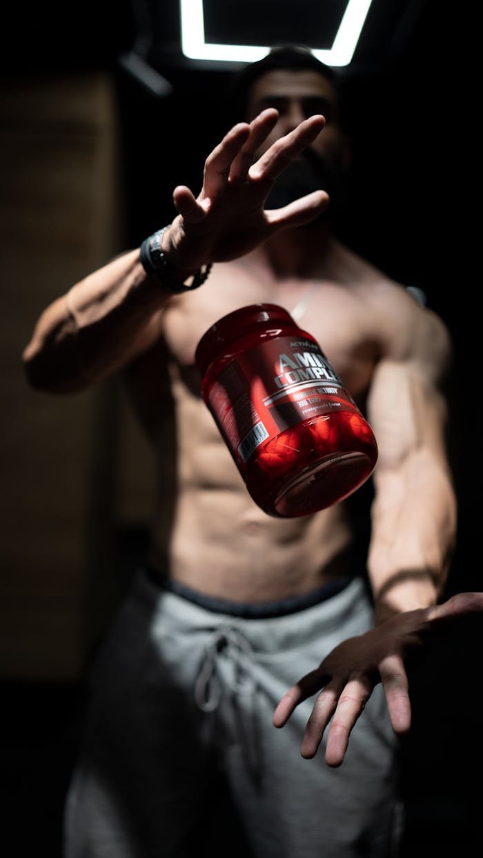 Services A shirtless man skillfully tosses a supplement jar in a dramatic indoor setting, showcasing fitness and agility.